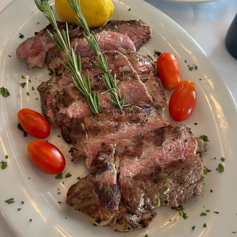 Sliced grilled steak with rosemary sprigs, cherry tomatoes, and a lemon wedge on a white plate.